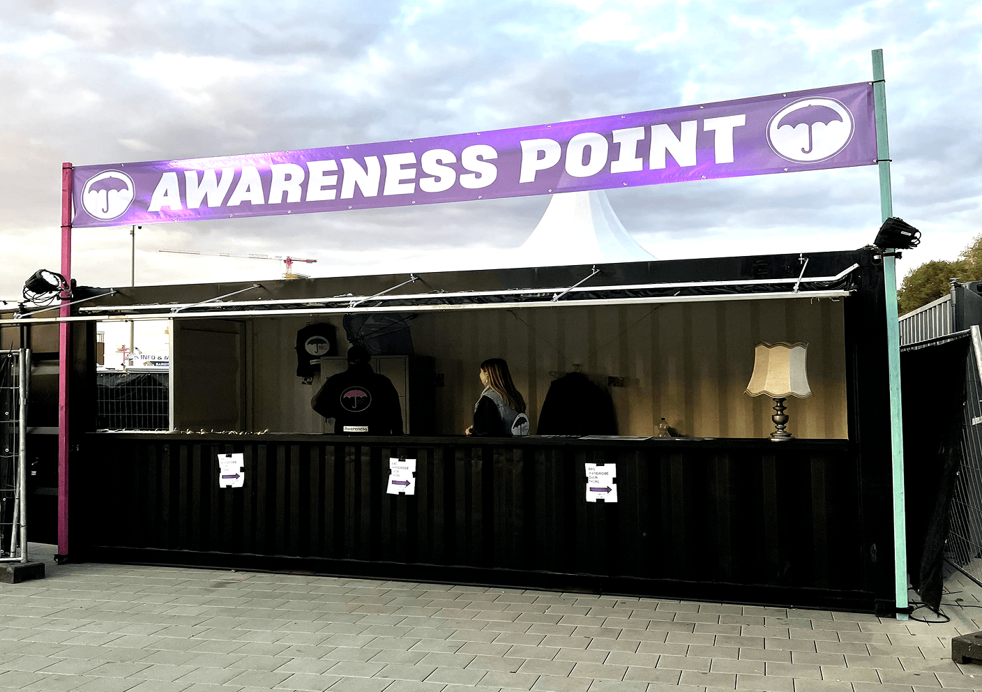 Schwarzer Infostand mit lila Banner „AWARENESS POINT“, zwei Personen und einer Tischlampe im Inneren.