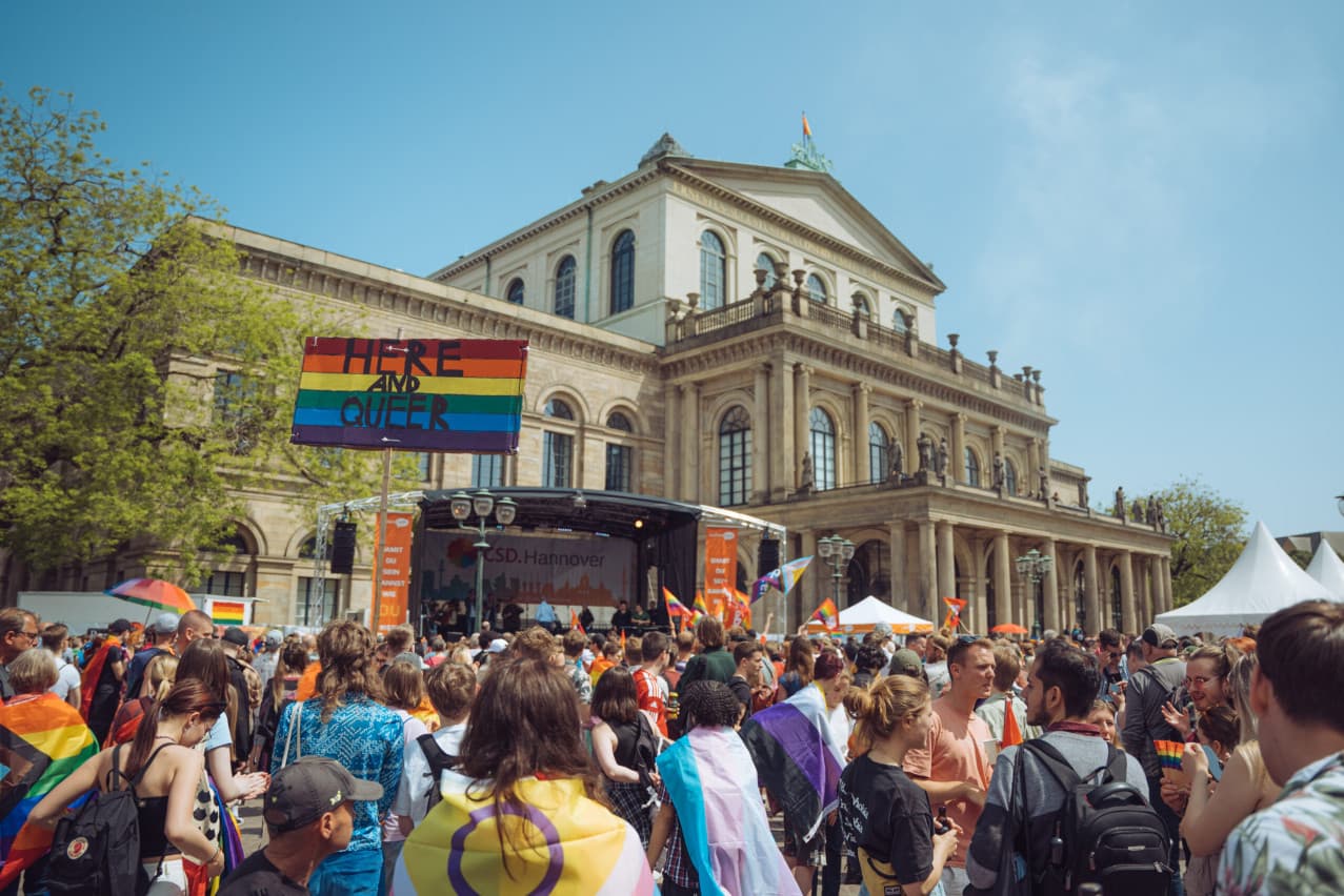 Pride-Demonstration vor dem Opernhaus Hannover mit Regenbogen-Schild „Here and queer“ und Fans mit Flaggen.