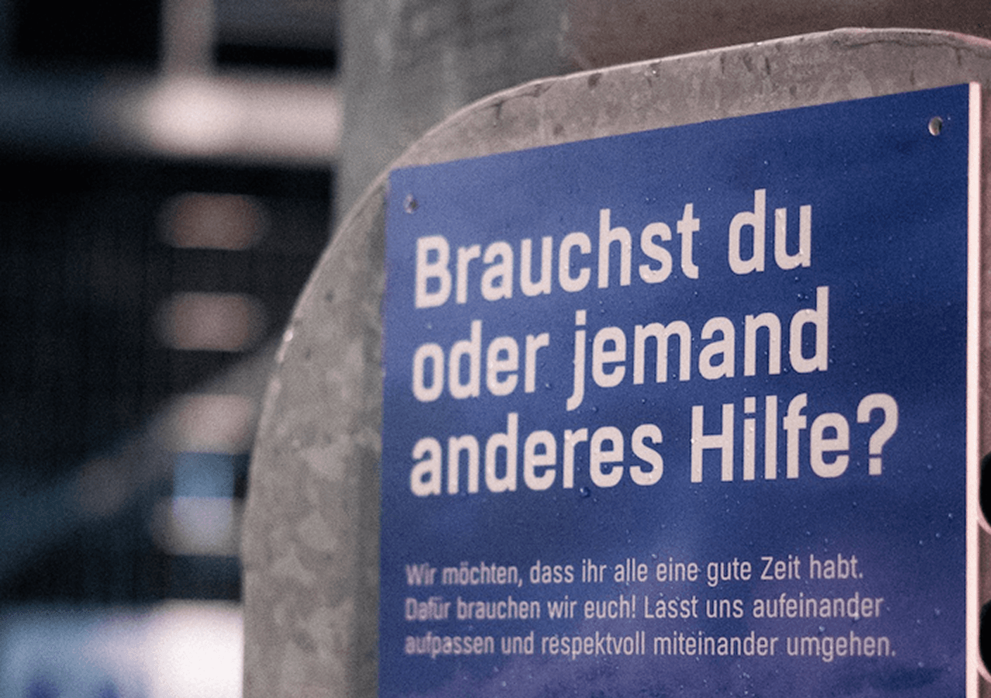 Blaues Schild mit der Aufschrift „Brauchst du oder jemand anderes Hilfe?“ und QR-Code im Stadion.