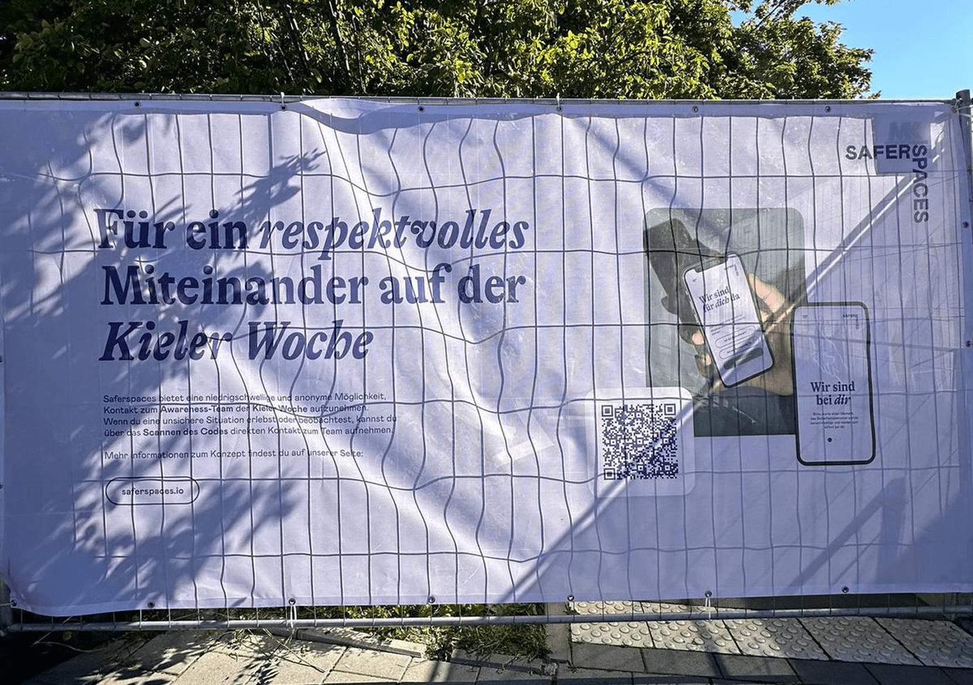 Banner mit Text: „Für ein respektvolles Miteinander auf der Kieler Woche“, QR-Code und Handy mit App „Wir sind bei dir!“.