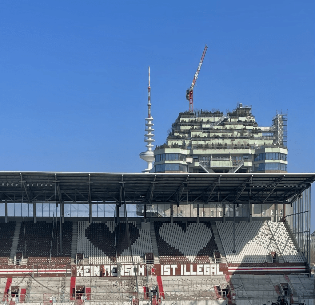 Tribüne des Millerntor-Stadions mit dem Schriftzug „Kein Mensch ist illegal“ und Großstadt-Hintergrund.