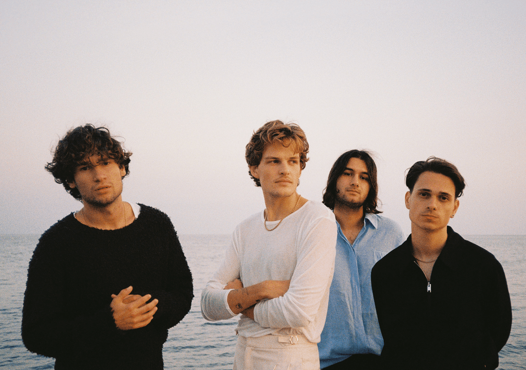 Four young men (Jeremias) standing next to each other at the sea at sunset, looking seriously into the camera.