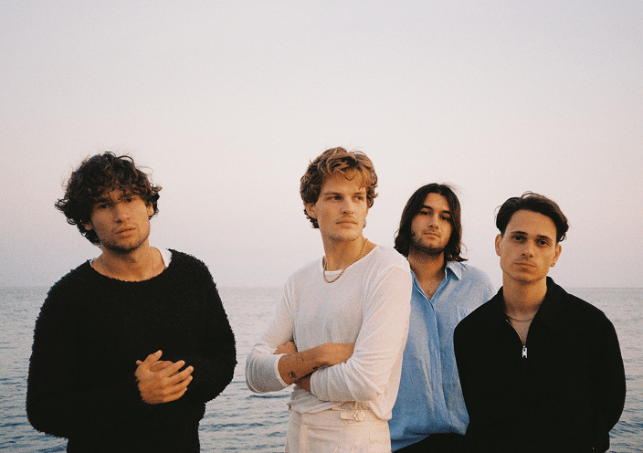 Four young men (Jeremias) standing next to each other at the sea at sunset, looking seriously into the camera.