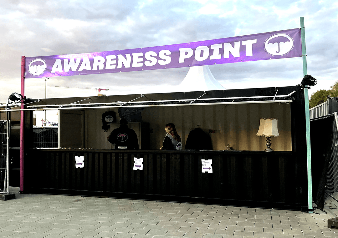 Black information stand with lavender banner „AWARENESS POINT“, two people and a table lamp inside.