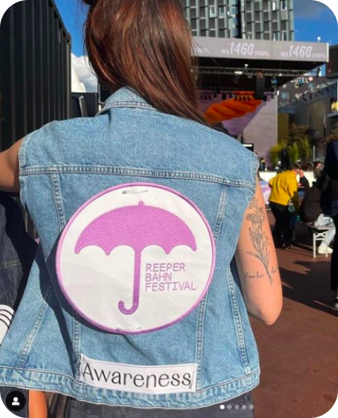 Eine Nahaufnahme des Rückens einer Person ist zu sehen. Diese trägt eine Jeansweste mit einem Aufnäher, auf dem ein lila Regenschirm, „Reeperbahn Festival“ und darunter „Awareness“ steht.
