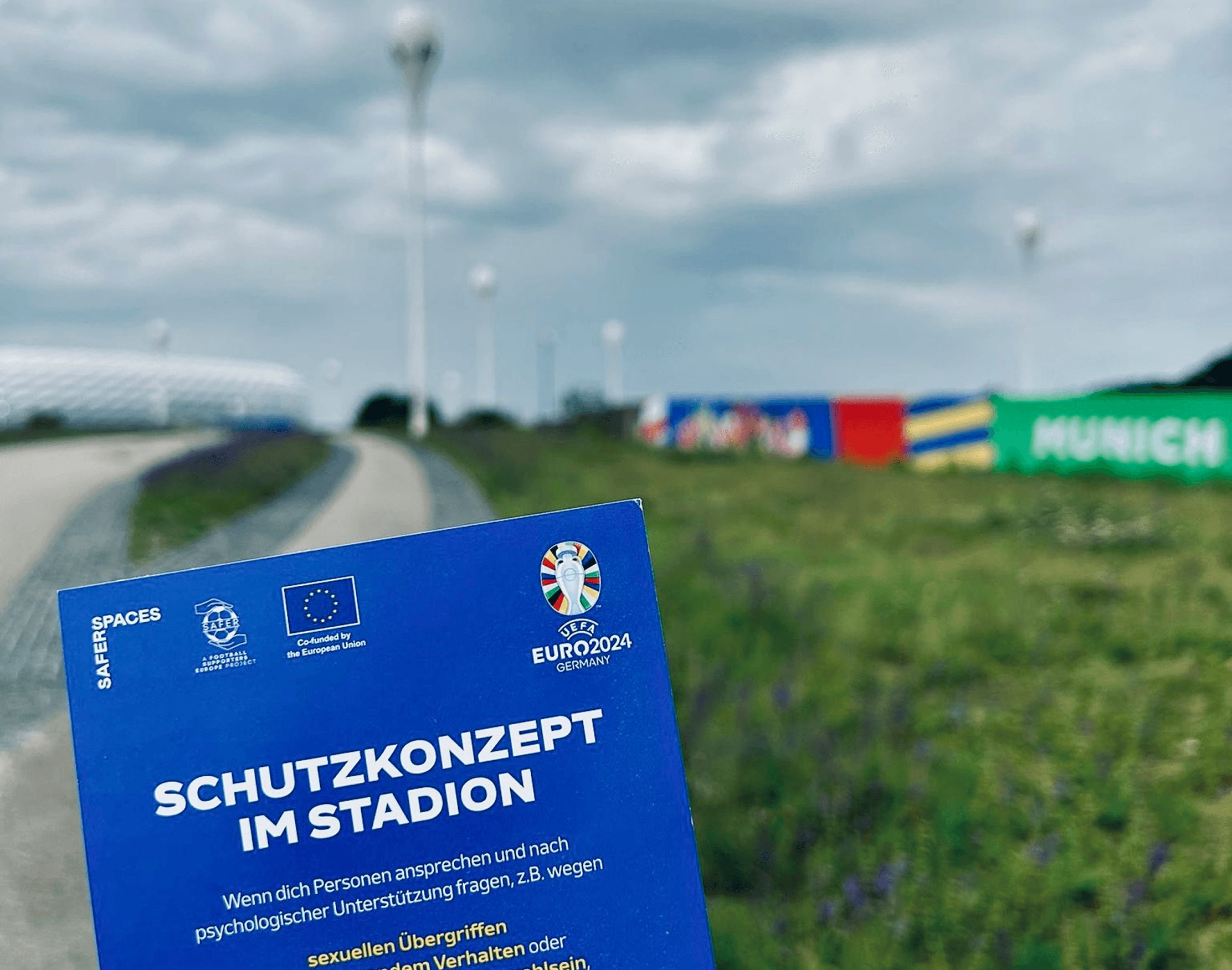 Blue flyer with „Schutzkonzept im Stadion“ in front of the stadium and colorful „Munich“ banner at cloudy sky.
